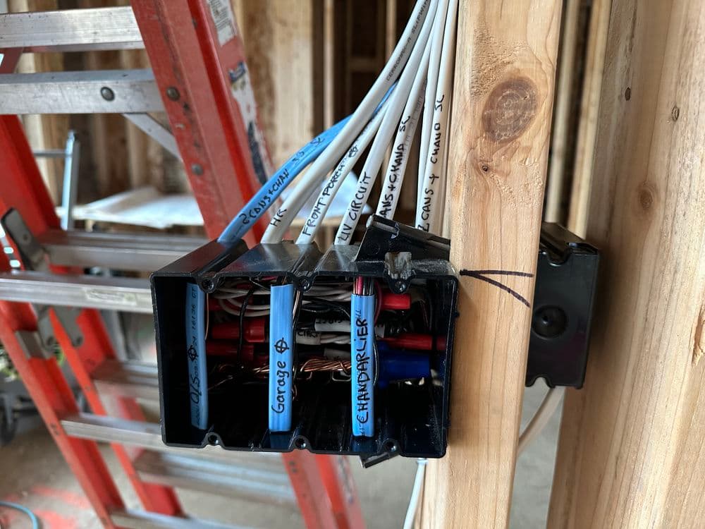 Electrical wiring box mounted on wooden studs with labeled wires in a construction setting.