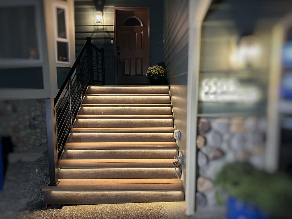 Well-lit outdoor stairs leading to a green house entrance with stone accents and flowers.