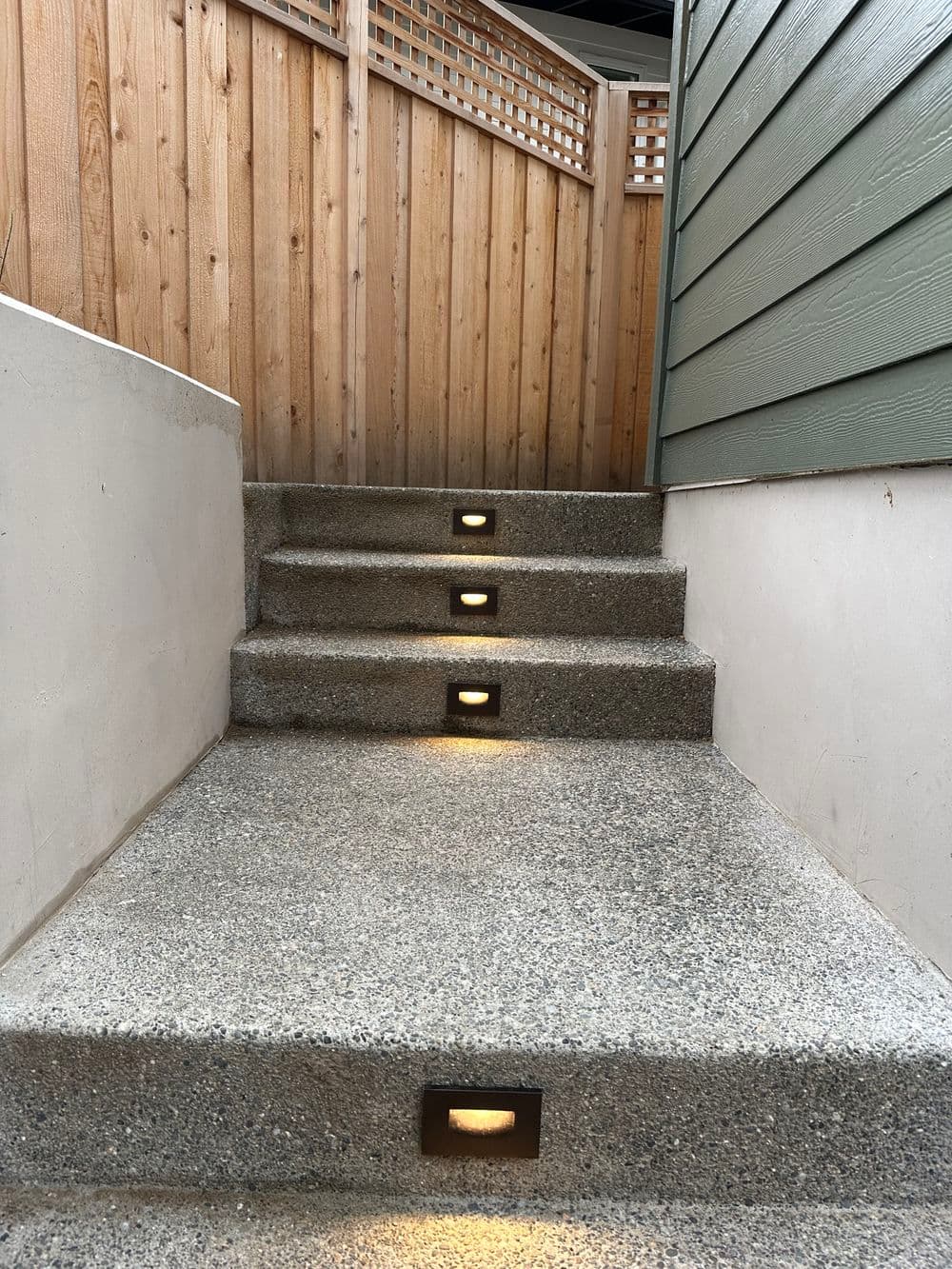 Modern stone staircase with recessed lighting leading to a wooden fence and green wall.